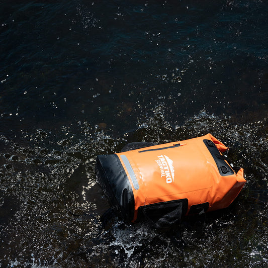 Bright orange Tactiko Survival dry bag partially submerged in dark, rippling water, with the white brand logo visible on the side.