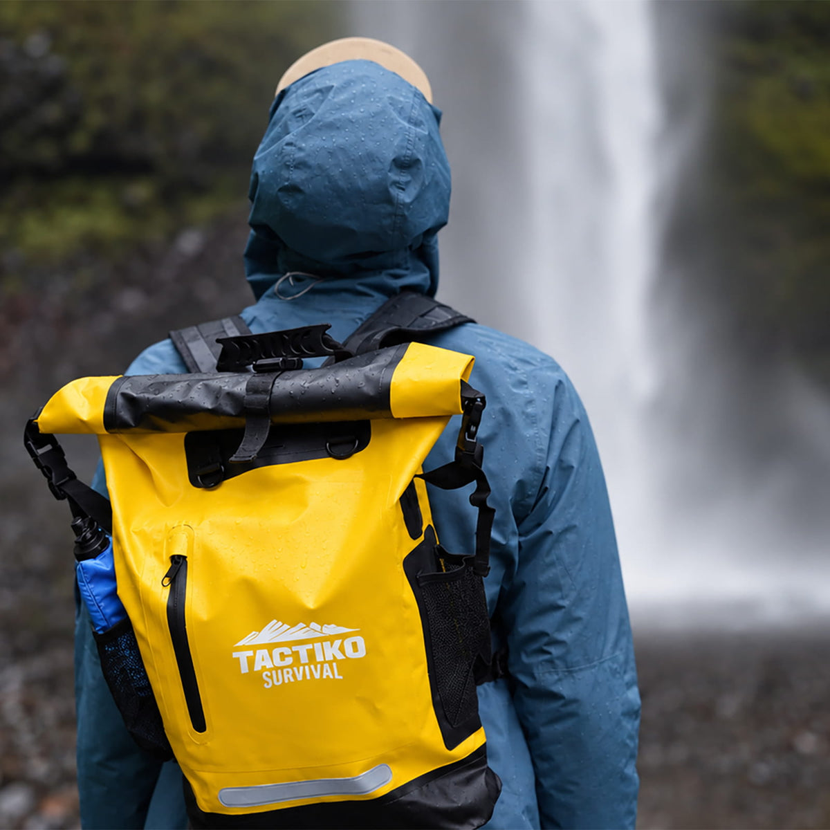 A person wearing a blue hooded rain jacket seen from behind, carrying a bright yellow TACTIKO SURVIVAL waterproof backpack with black straps and trim in front of a blurred waterfall background.